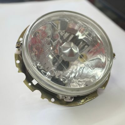 It features a clear lens with a multi-surface reflector design. The assembly includes a metal mounting or adjustment bracket. The headlight is compatible with a halogen H4 bulb type. It is often used as an aftermarket upgrade for older vehicles.